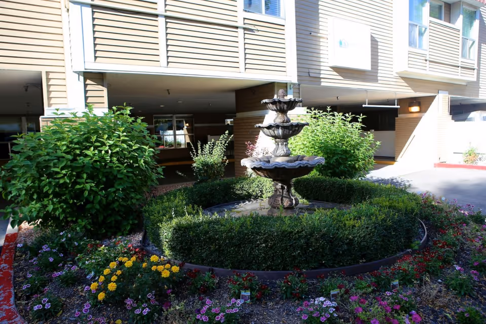 Outdoor garden area with a three-tiered stone fountain surrounded by neatly trimmed bushes and colorful flowers in front of a beige building with windows and an open parking area underneath.