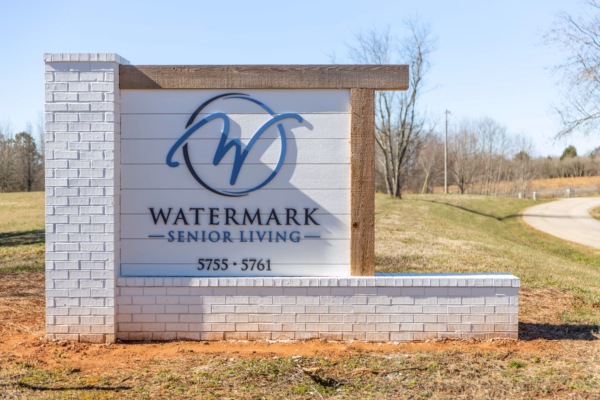 A freestanding white brick entrance sign with a blue circular 'W' logo that reads 'Watermark Senior Living' beside a curved roadway.
