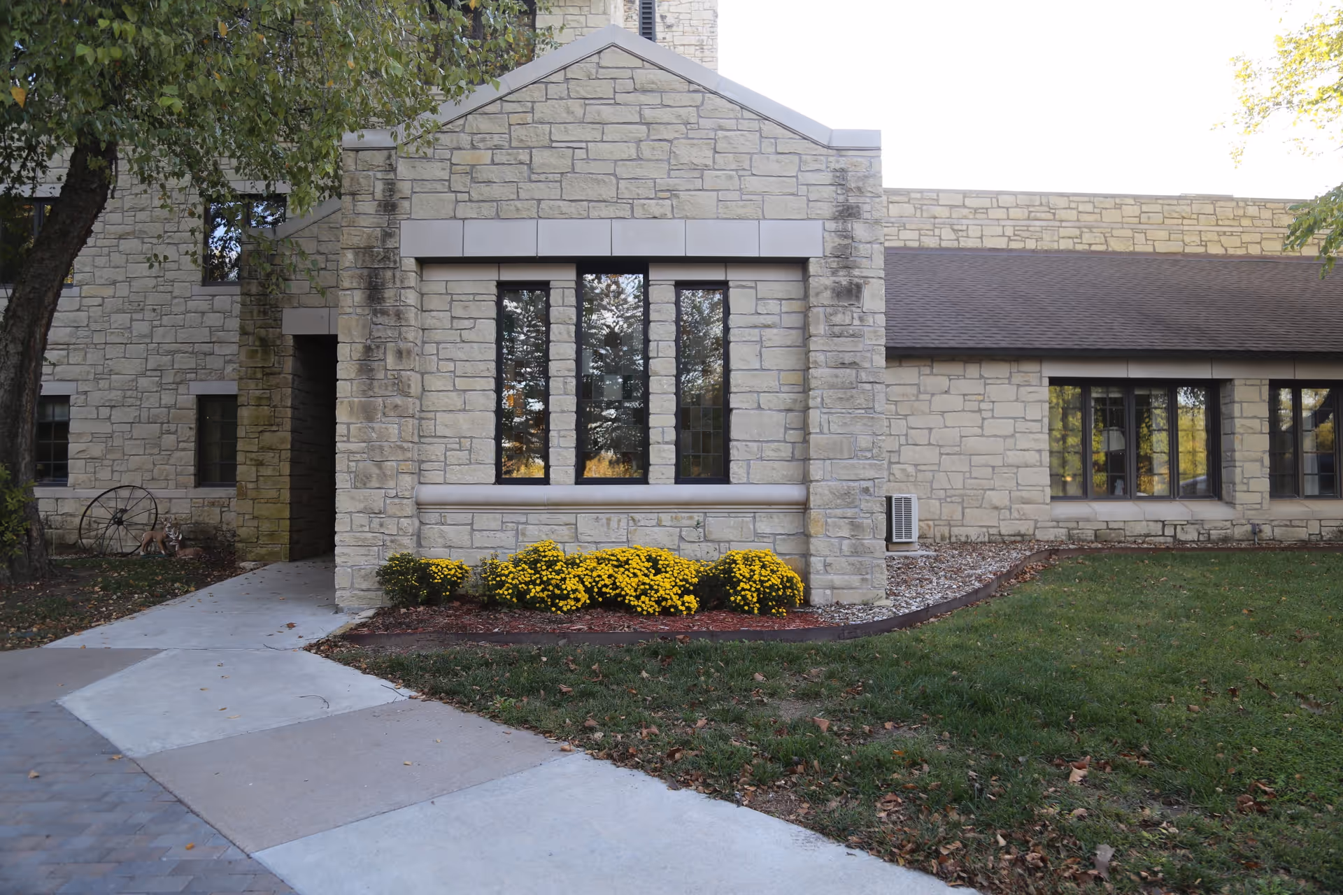 Stone-faced building front with tall windows, a bed of yellow flowers, and a curved concrete walkway.