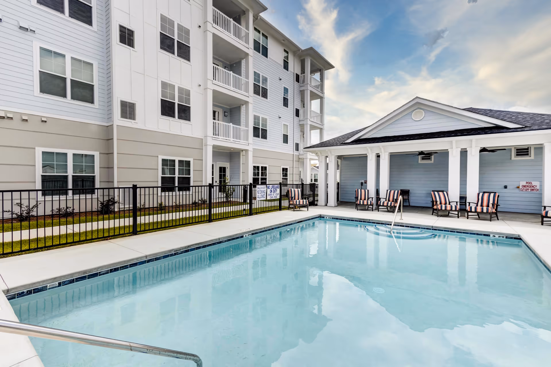Outdoor swimming pool and patio with lounge chairs beside a multi-story residential building under a partly cloudy sky.