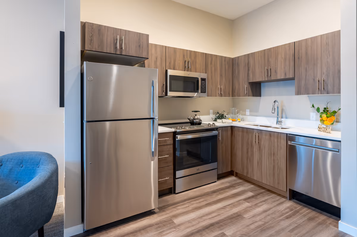 Modern kitchen with stainless steel refrigerator, oven, microwave, and dishwasher. The kitchen features wood cabinets, a white countertop, a sink with a faucet, and a small plant decoration on the counter. The floor has wood-like vinyl planks.