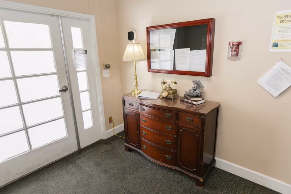 Interior view of a senior living facility entrance area with a wooden sideboard holding a vintage telephone, a lamp, and a small statue. A bulletin board with papers is mounted on the wall above the sideboard. A glass-paneled door is on the left side, and a fire alarm is visible on the wall.