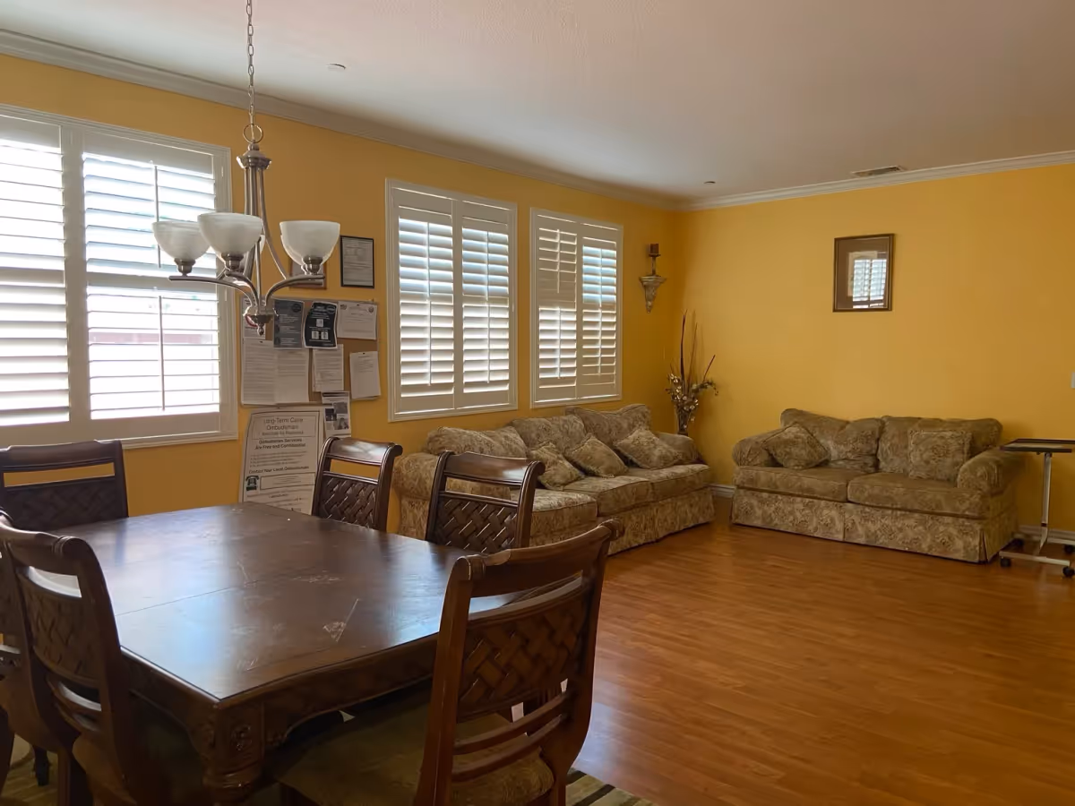 Sunlit common room with a wooden dining table and chairs in the foreground and two patterned sofas against a yellow wall.
