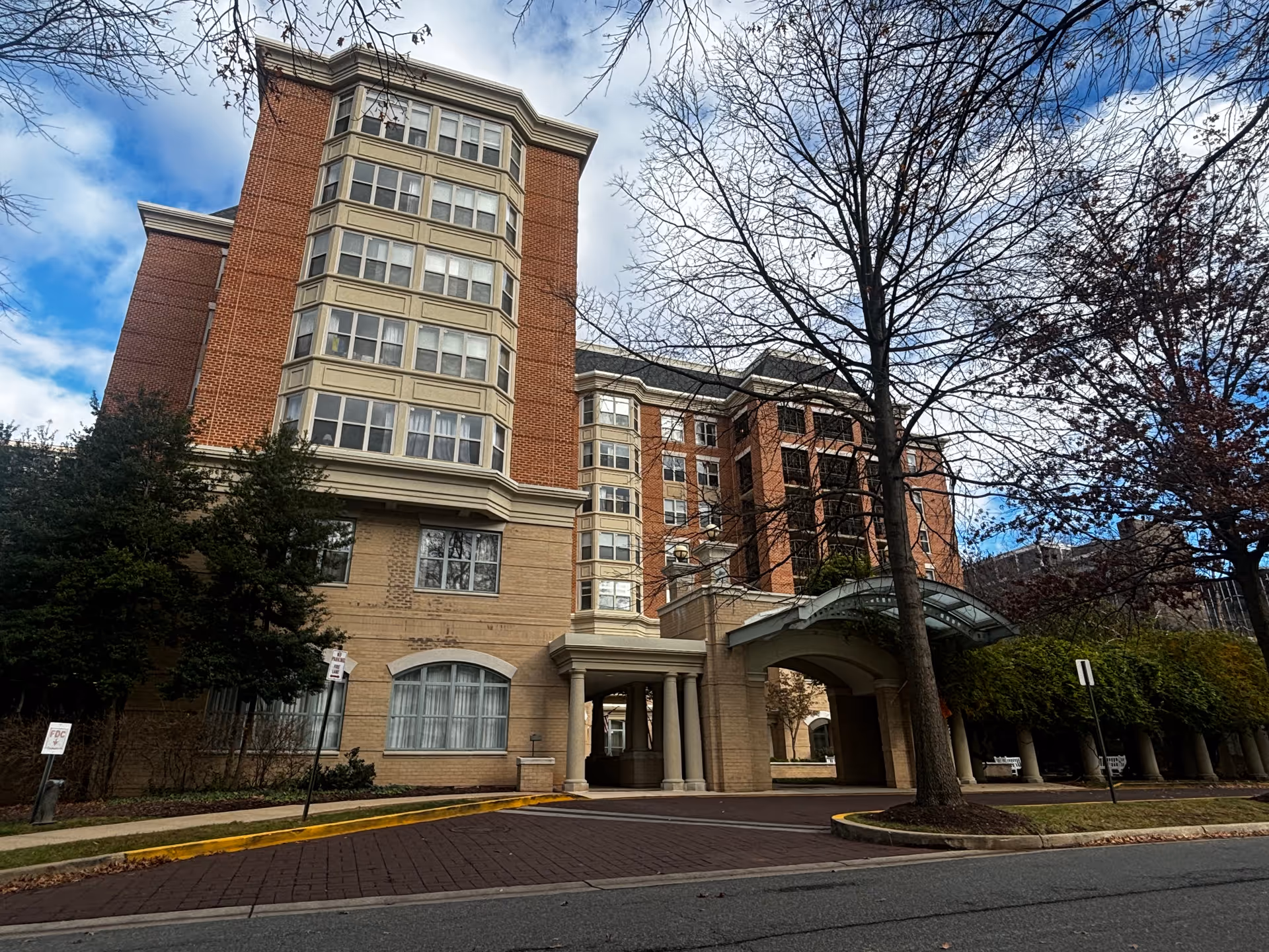 Exterior view of a multi-story brick building with large windows and a covered entrance driveway. Trees with bare branches and some greenery surround the building under a partly cloudy sky.