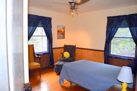 Bedroom with a blue-covered bed, two windows with dark blue curtains, wood-paneled lower walls, a chair with a yellow pillow, and a small bedside lamp.