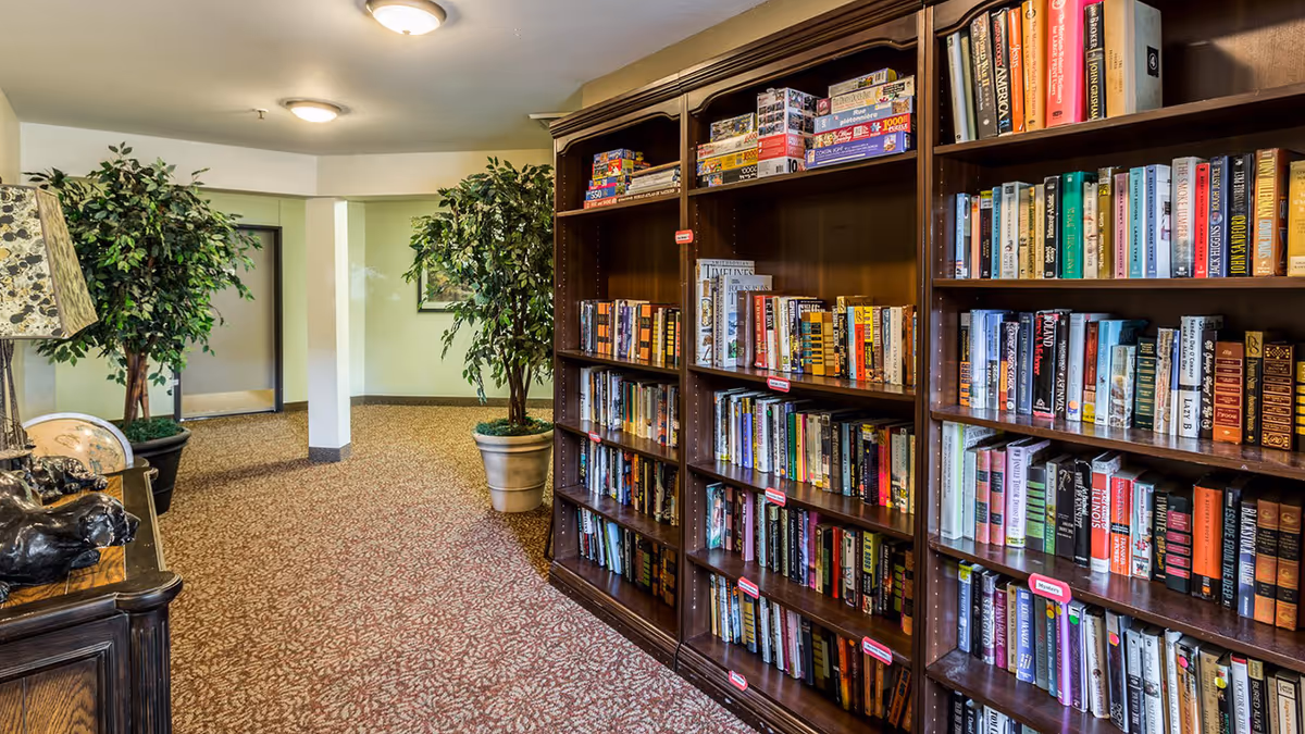 Interior view of a senior living facility hallway with carpeted floor, two large potted plants, a wooden bookshelf filled with books and board games, and a decorative table with a lamp and globe.