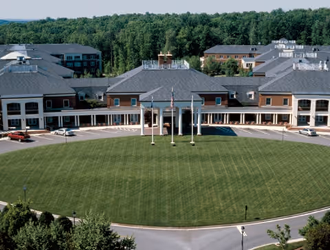A large senior living facility building with a circular driveway and a well-maintained lawn in front. The building has multiple sections with gray roofs and brick walls, surrounded by trees in the background.