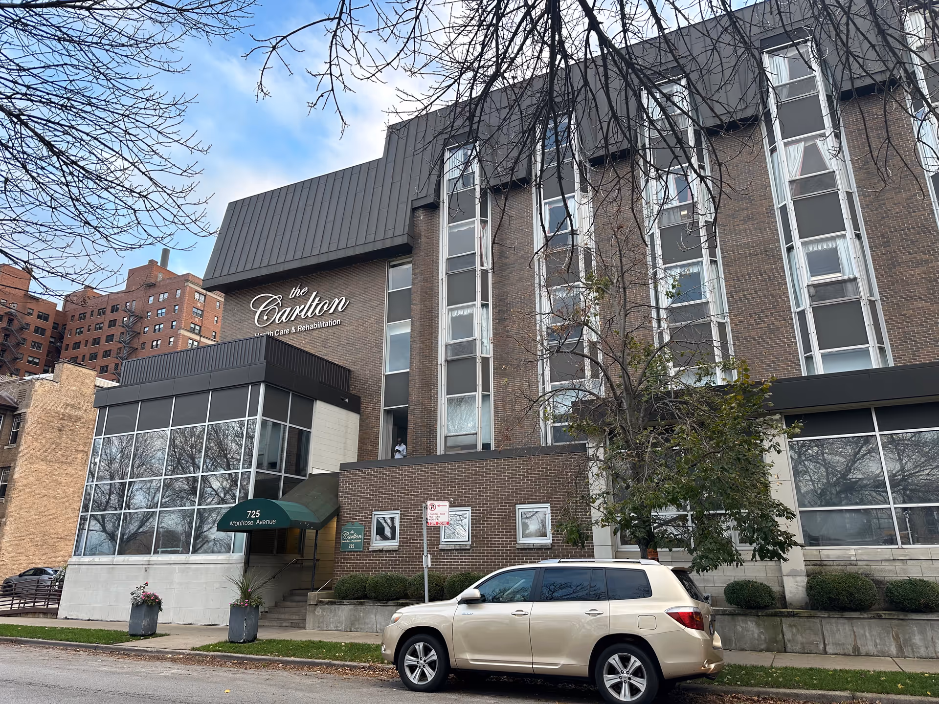 Exterior view of a multi-story brick building with large windows and a green awning over the entrance. The building has a sign that reads 'the Carlton Health Care & Rehabilitation' and the address '725 Montrose Avenue' is visible. A beige SUV is parked on the street in front of the building, and leafless tree branches frame the top of the image.