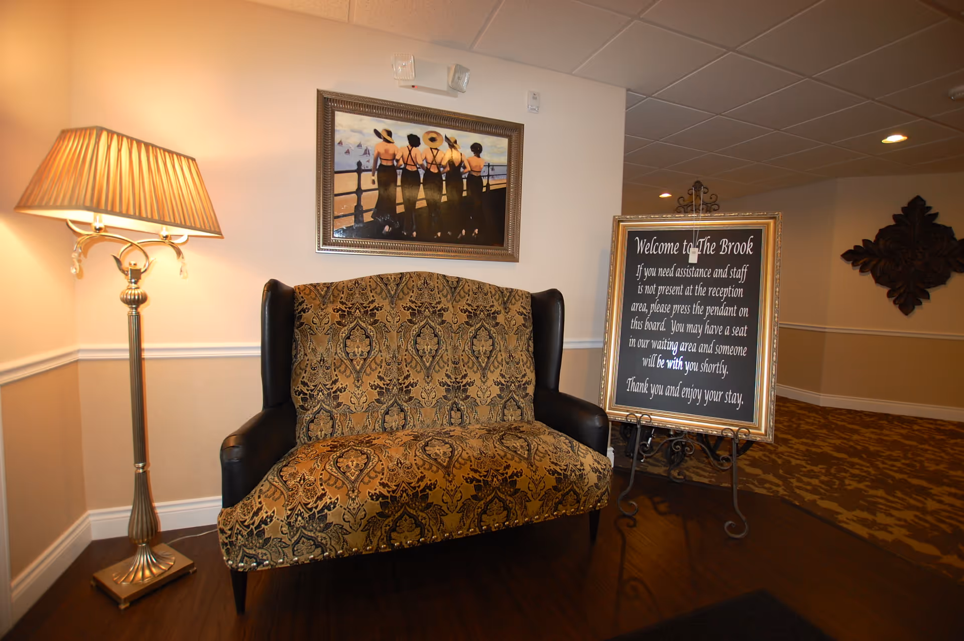 A decorated seating area with a patterned loveseat, floor lamp, framed artwork, and a framed welcome sign on an easel in a facility lobby.