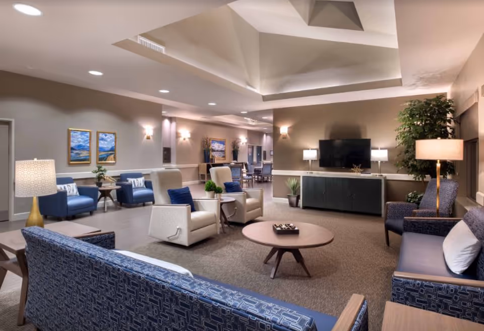 A spacious and well-lit living room area in a senior living facility with multiple seating options including blue patterned sofas, beige recliners with blue pillows, and blue armchairs. The room features a large flat-screen TV mounted on the wall above a dark cabinet, several lamps, potted plants, and framed landscape paintings on the walls. The ceiling has a recessed design with soft lighting, and a dining area is visible in the background.