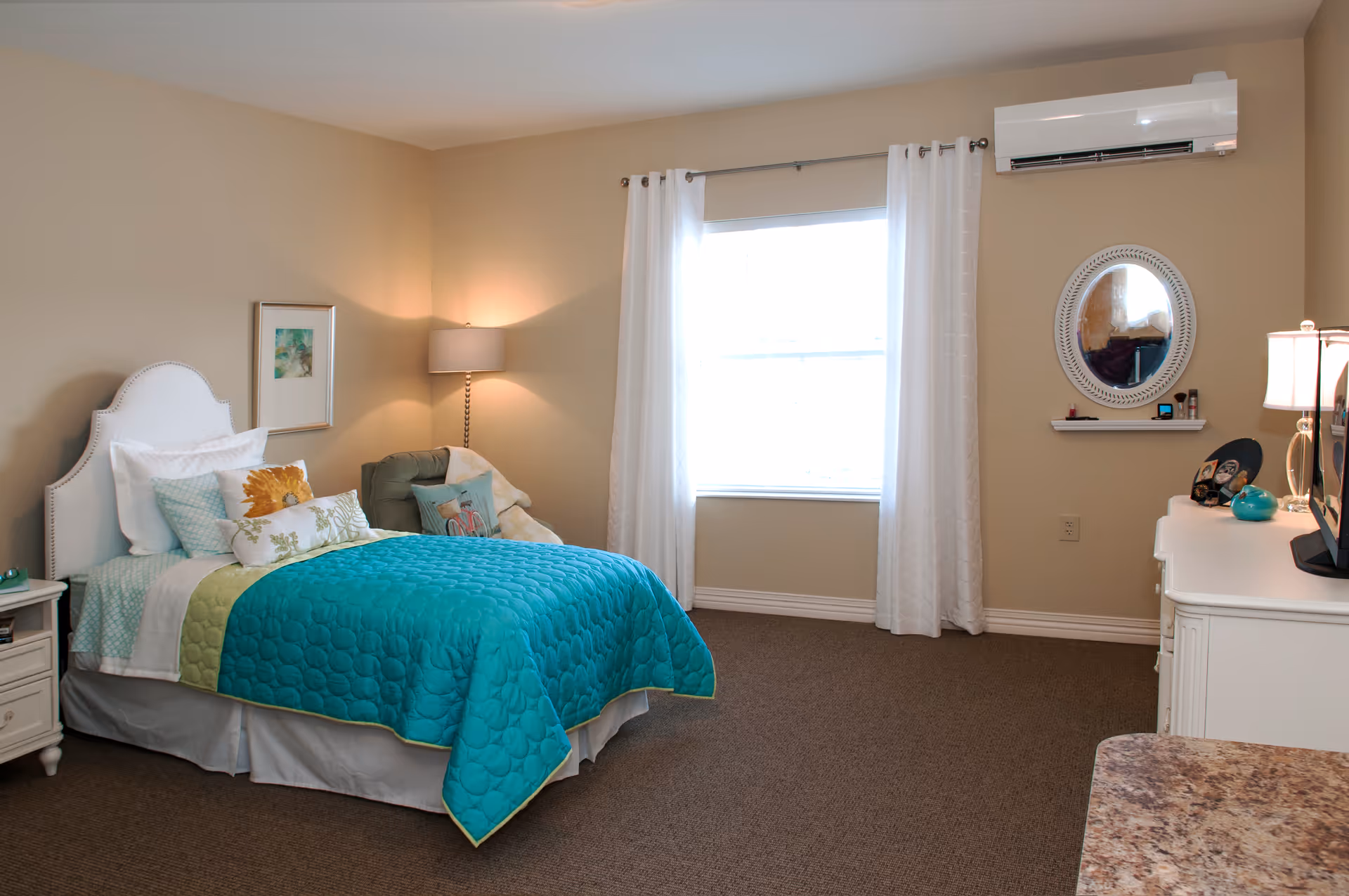 A tidy bedroom with a bed dressed in a turquoise quilt, nightstand, lamp, window with white curtains and a dresser with mirror.