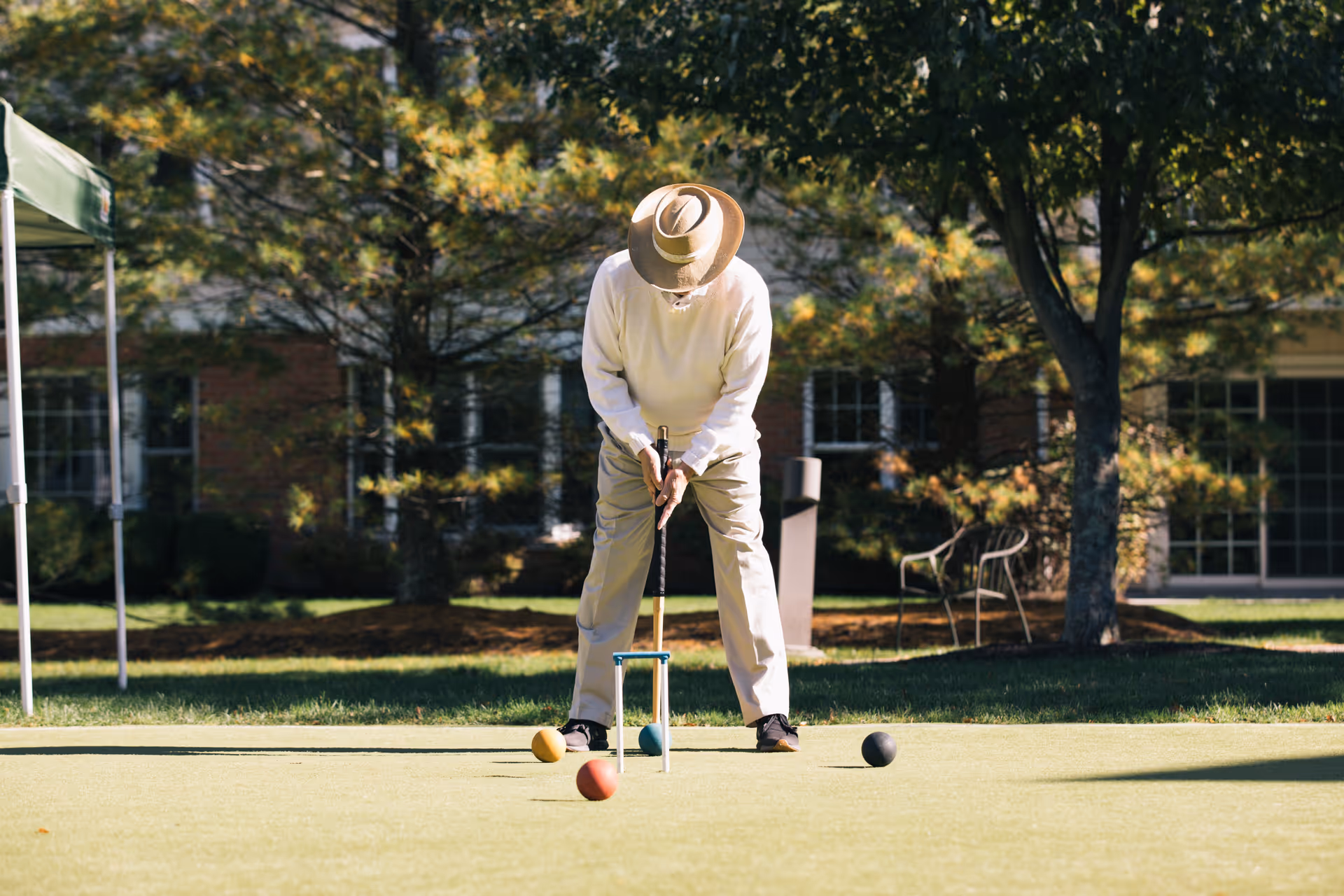 An elderly person wearing a hat, white sweater, and beige pants is playing croquet on a grassy lawn with trees and a building in the background.