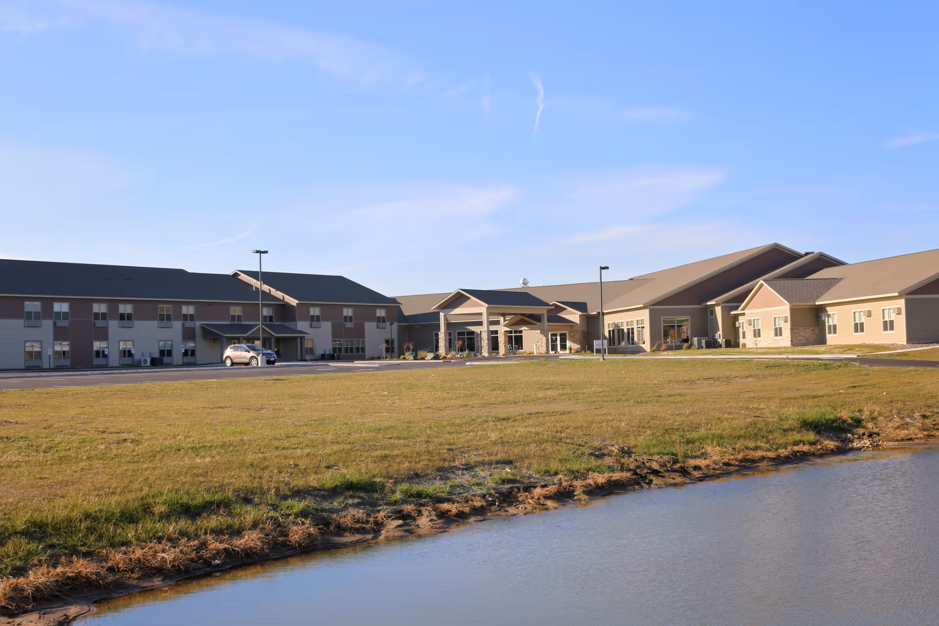 Exterior front view of a low-rise senior living facility by a grassy lawn and small pond under a blue sky.