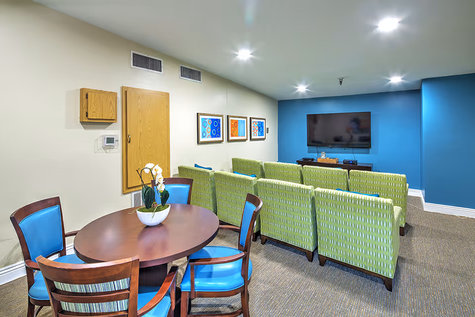 A cozy senior living common area with a round wooden table and four chairs with blue cushions in the foreground. Behind the table, there are two rows of green patterned armchairs facing a wall-mounted flat-screen TV on a blue accent wall. Three colorful framed artworks hang on the adjacent beige wall.