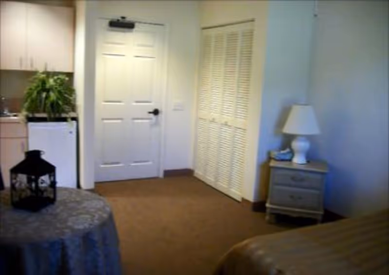 Small studio-style room showing a bed corner and nightstand with lamp, a round table, a kitchenette with mini-fridge and plant, and a door with closet doors.