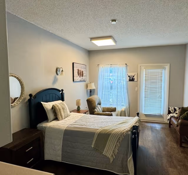 Furnished bedroom with a bed, nightstands, an armchair, and a window with sheer curtains overlooking a door with blinds.