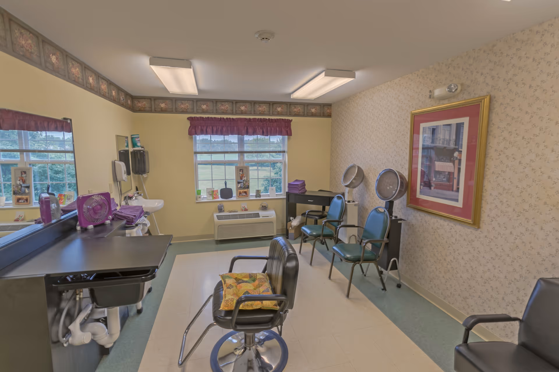 Interior view of a hair salon area in a senior living facility with a black salon chair in the center, two blue chairs with hair dryers on stands against the wall, a sink with a mirror and hair care items on the left, and a window with purple valance letting in natural light.