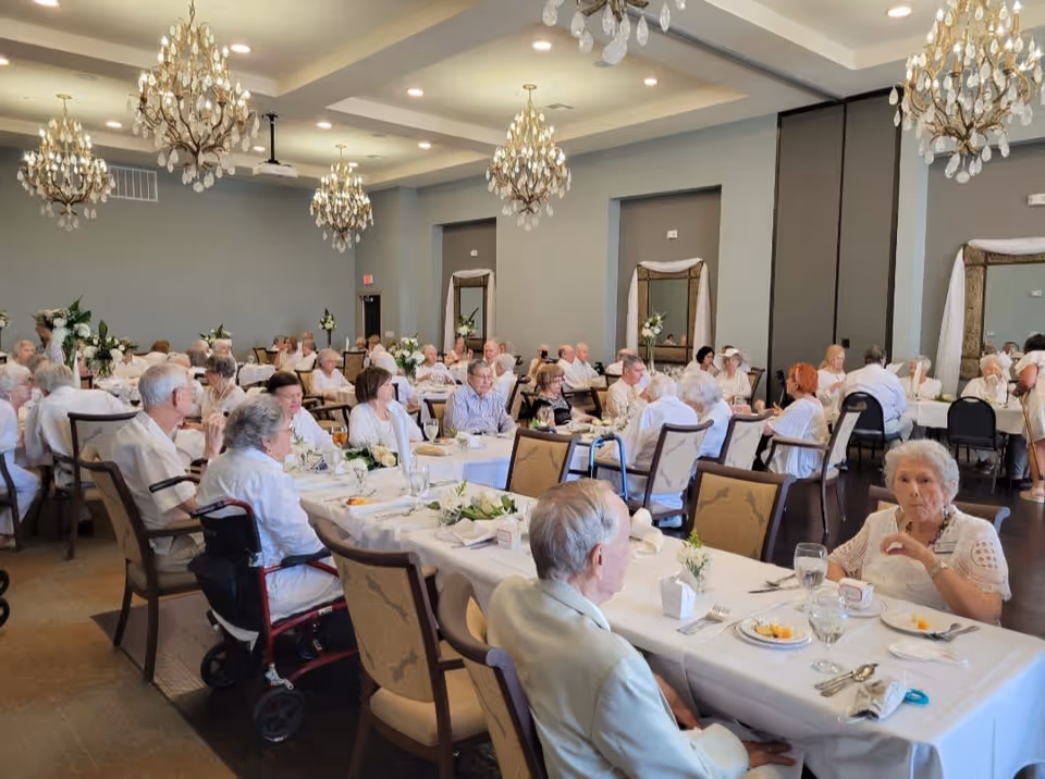 A large dining room filled with elderly people seated at tables covered with white tablecloths, enjoying a meal together. The room has elegant chandeliers hanging from the ceiling and large mirrors on the walls. Some individuals are using walkers or wheelchairs.
