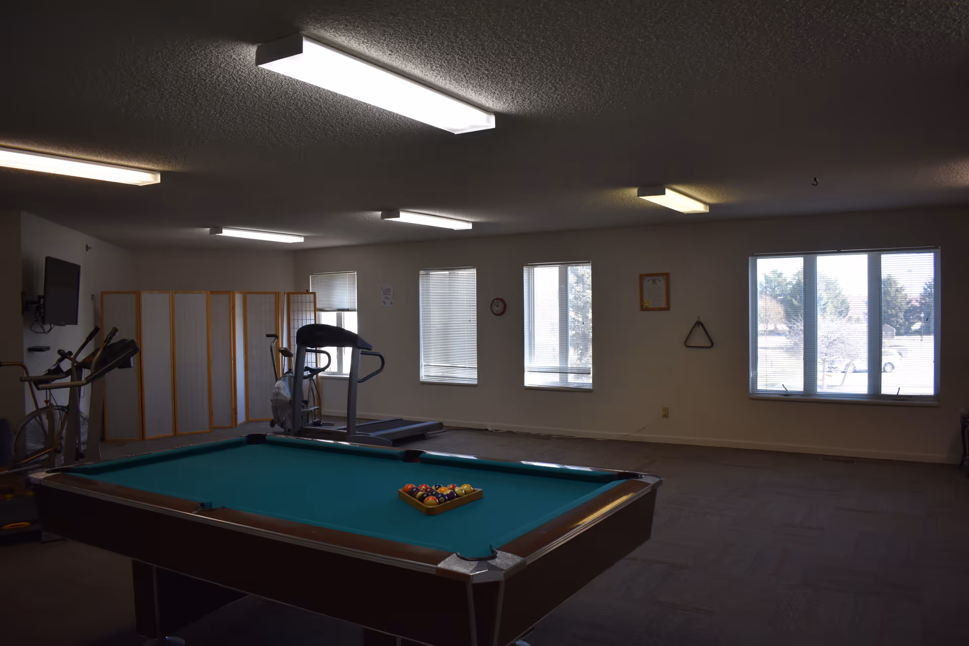 Recreation room with a pool table in the foreground and exercise equipment near windows along the far wall.