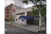 Exterior view of Brooklyn Center For Rehab And Residential Health Care, showing a white building with a blue awning over the entrance. There are trees and a sidewalk in front, with a street and parked cars visible.