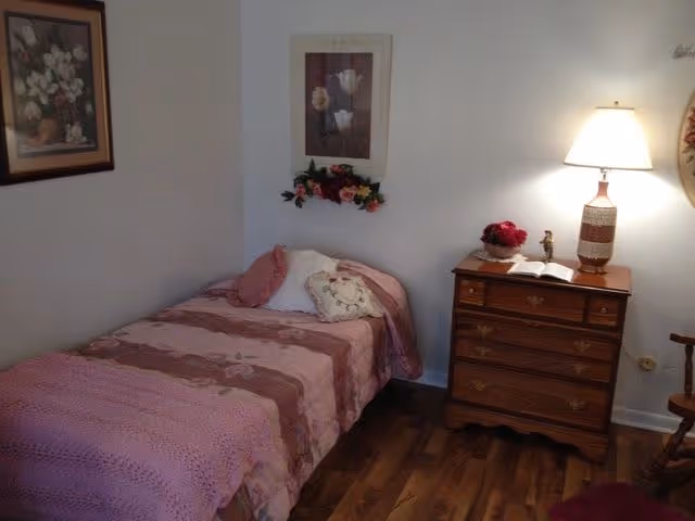A small bedroom with a single bed covered in a pink and mauve patterned bedspread and pillows. Next to the bed is a wooden dresser with a lamp, a small flower arrangement, and an open book. The walls are decorated with framed floral artwork and a small floral wreath. The floor is wooden.