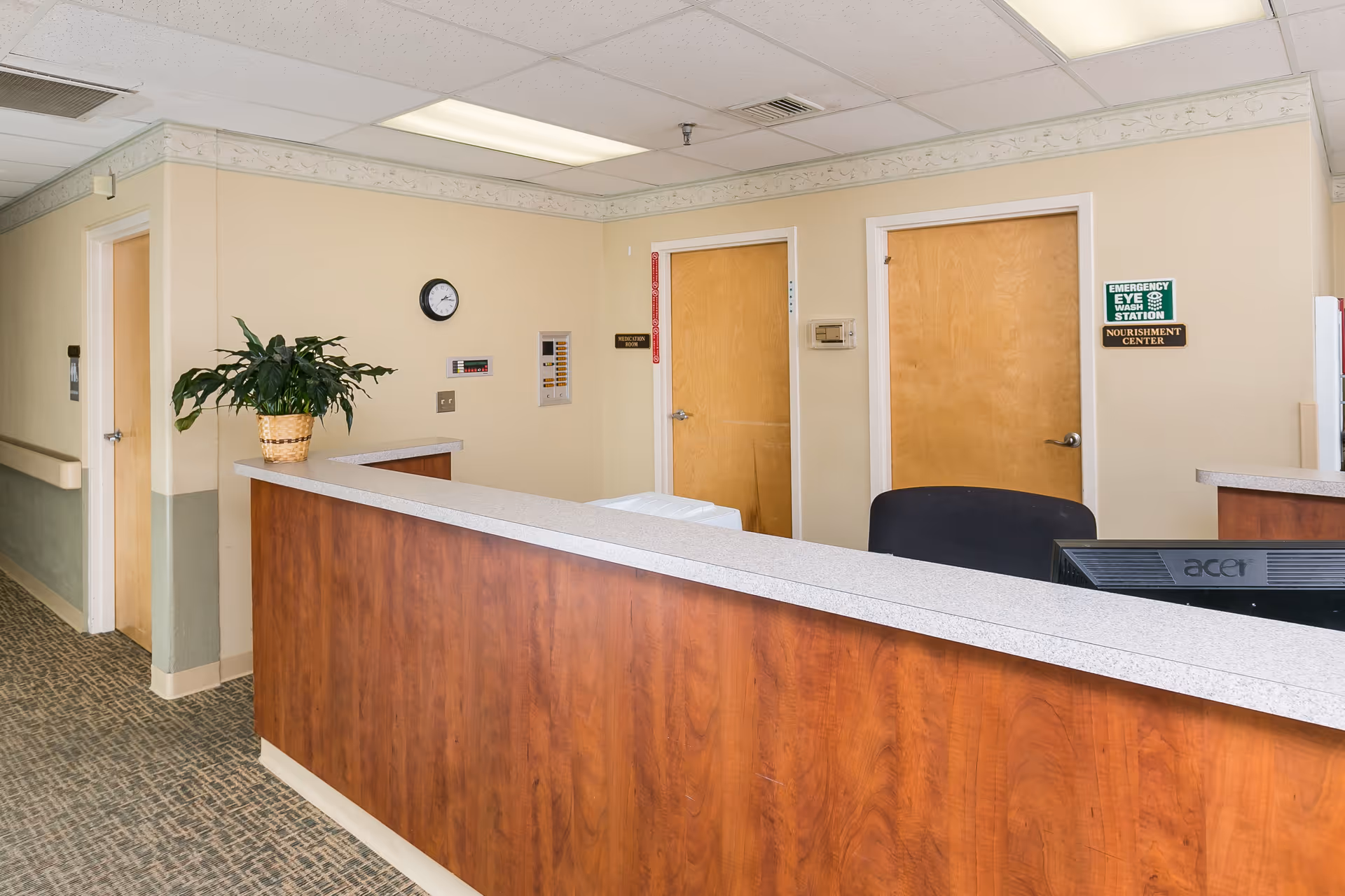 Reception desk area inside a nursing facility with a wood counter, potted plant, office chair, and several closed doors.