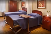Shared senior care room with two adjustable beds covered in blue linens, wooden nightstand and wardrobe, framed artwork and hardwood flooring.
