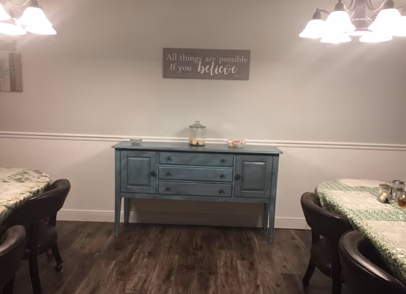 Interior view of a dining area with a blue sideboard against a white wall. On the wall above the sideboard is a sign that reads 'All things are possible if you believe.' Two tables with green and white tablecloths and dark chairs are partially visible on either side. The floor is wooden, and ceiling lights are visible at the top corners.