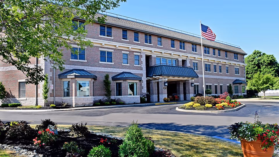 Front exterior view of a three-story brick building with multiple windows, a covered entrance, an American flag on a flagpole, and landscaped flower beds and shrubs in front under a clear blue sky.