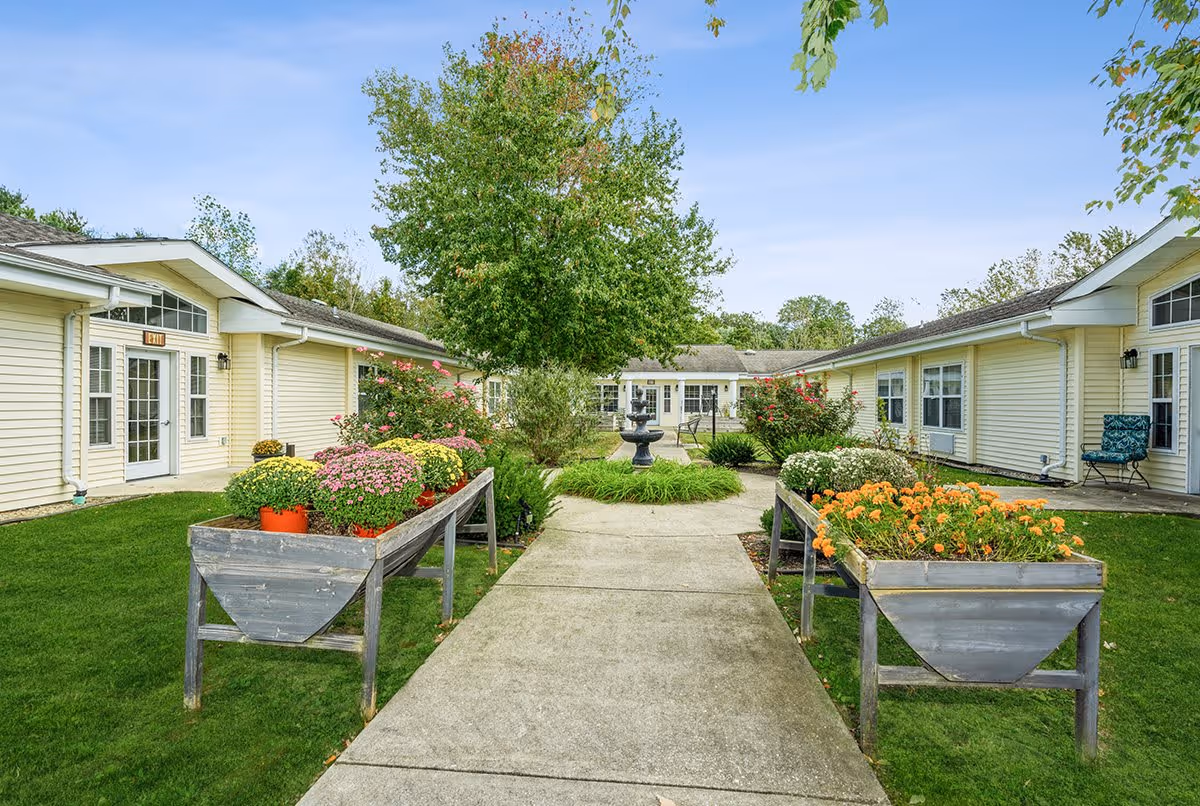 Outdoor courtyard area at Cedar Creek of Bedford with a concrete walkway flanked by raised flower beds filled with colorful flowers. There are light yellow buildings on both sides with windows and doors, a tree in the center background, and a black water fountain surrounded by greenery.