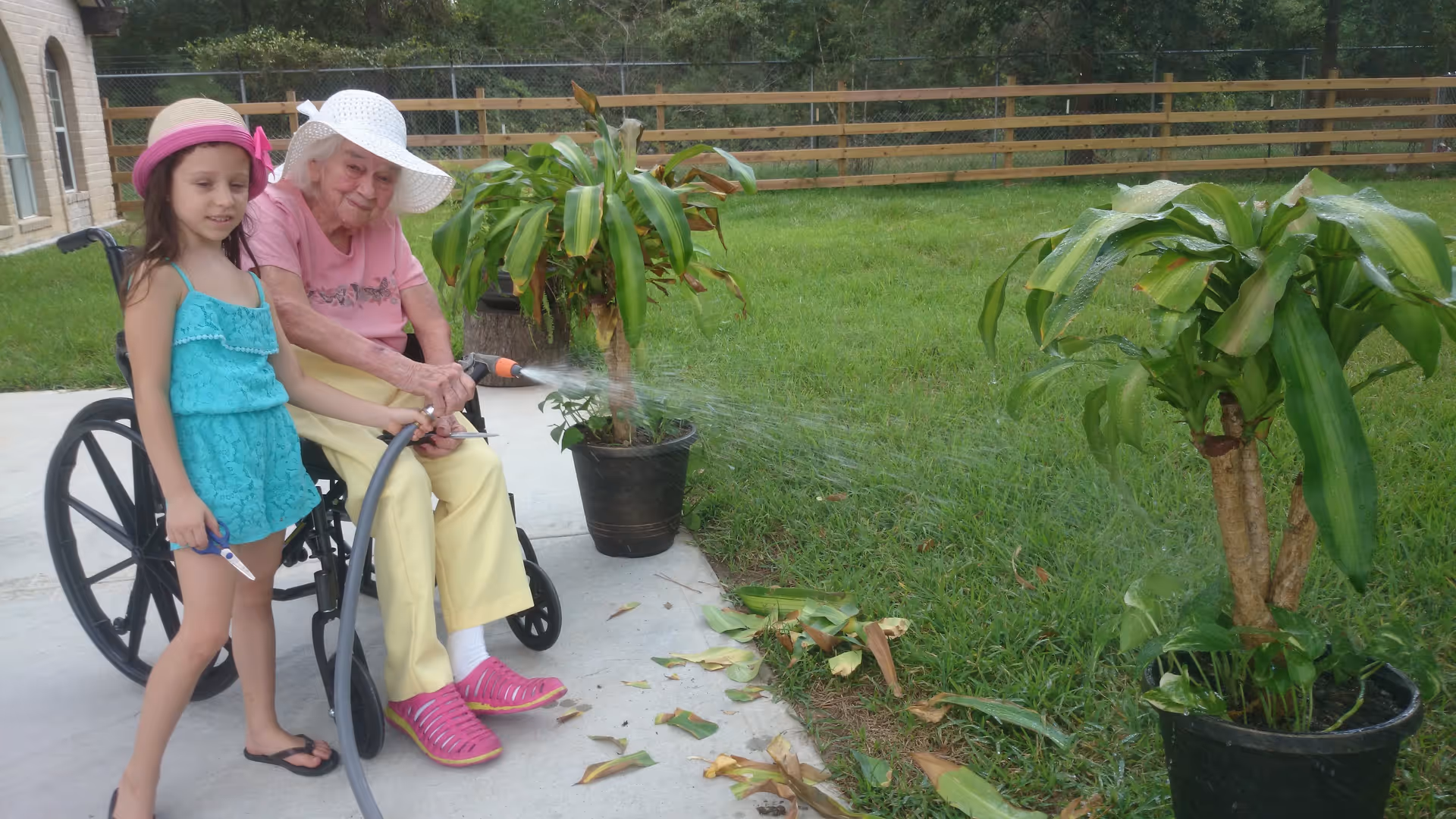 An elderly woman in a wheelchair wearing a white sunhat and pink shirt waters plants with a garden hose while a young girl in a turquoise dress and pink sunhat stands beside her holding the hose. They are outside on a concrete patio next to green grass and potted plants, with a wooden fence and trees in the background.