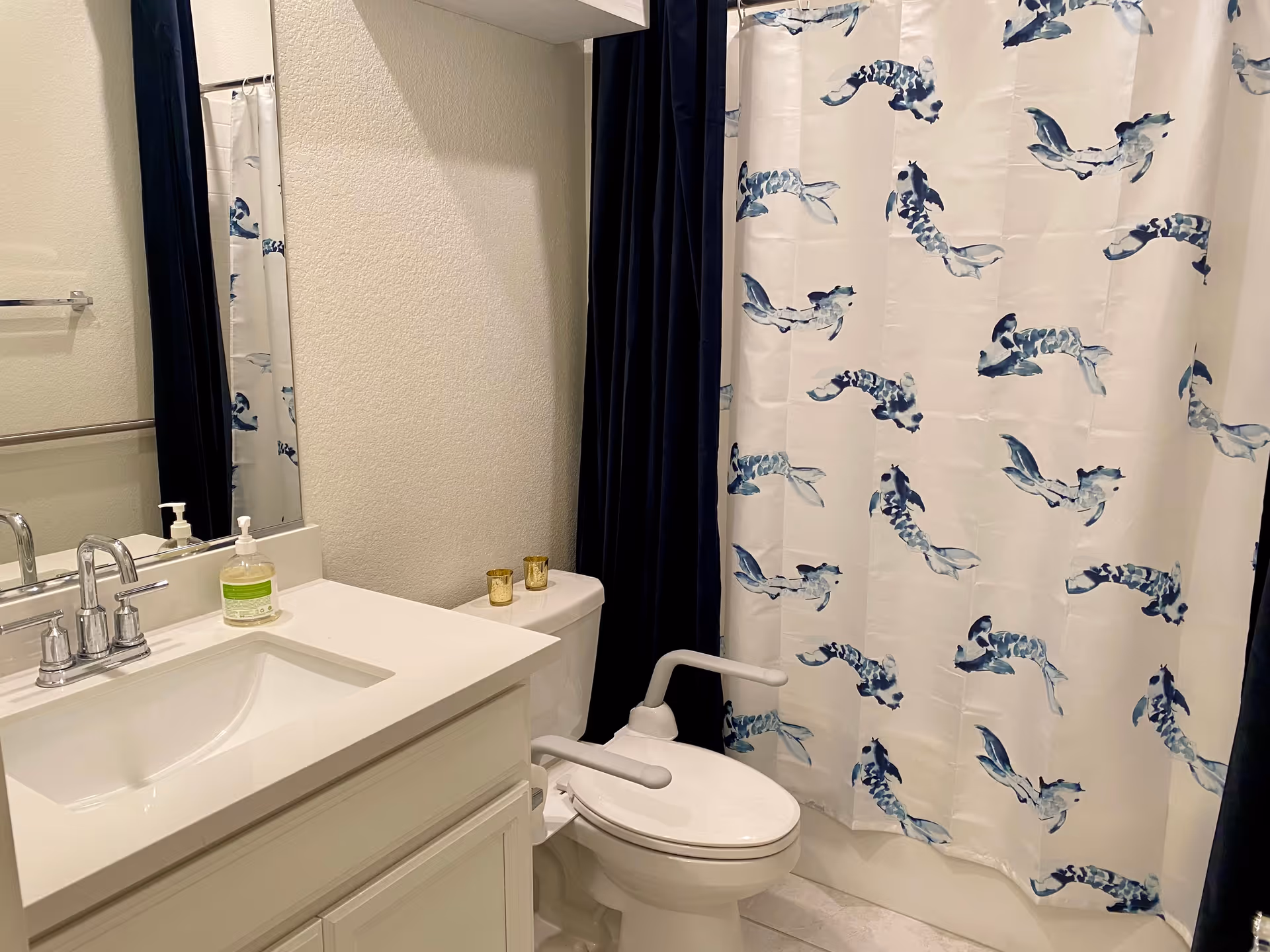 A clean bathroom with a white vanity and sink, a toilet fitted with safety rails, and a shower curtain patterned with blue fish.