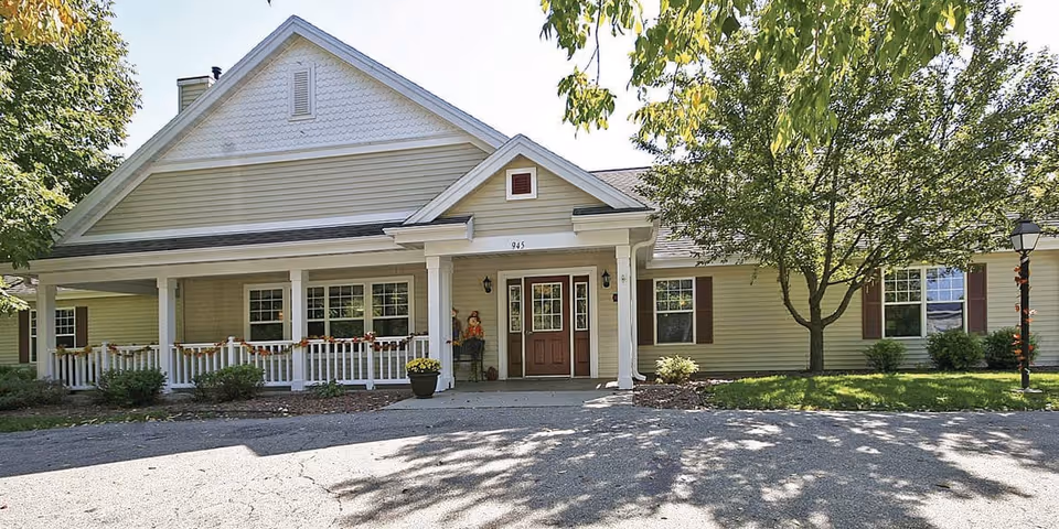 Exterior view of a single-story senior living facility building with beige siding, white trim, and a covered porch decorated with fall-themed garlands. There are trees and shrubs around the building, and a paved driveway in front.