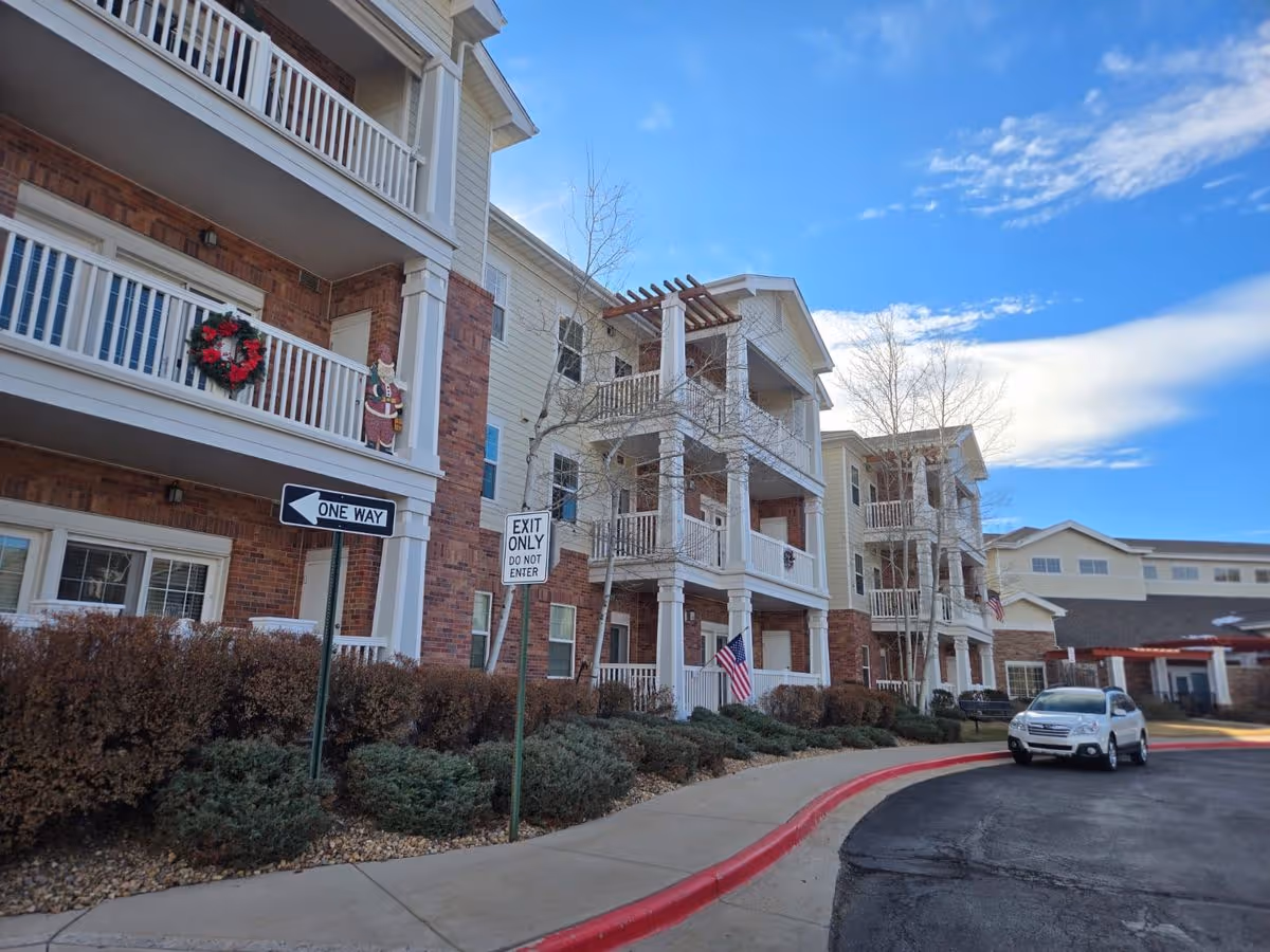Front exterior of a multi-story senior living building with balconies, American flags, and a curved driveway with a parked car.