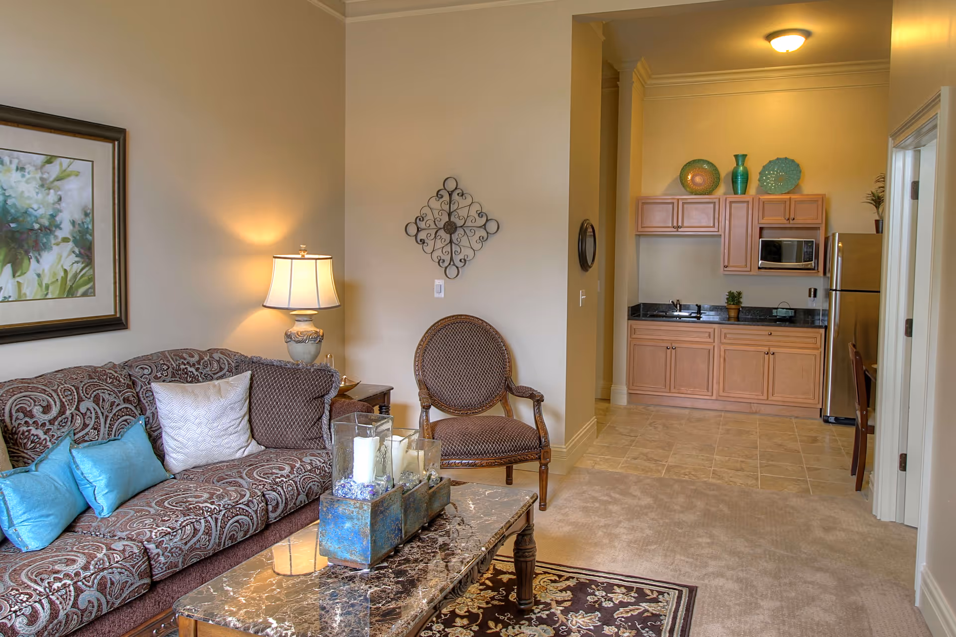 A cozy living area with a patterned sofa adorned with blue and white pillows, a wooden armchair, and a marble-top coffee table with decorative candles. A table lamp and framed floral artwork are on the wall behind the sofa. In the background, there is a small kitchenette with wooden cabinets, a microwave, a refrigerator, and decorative vases on top of the cabinets.