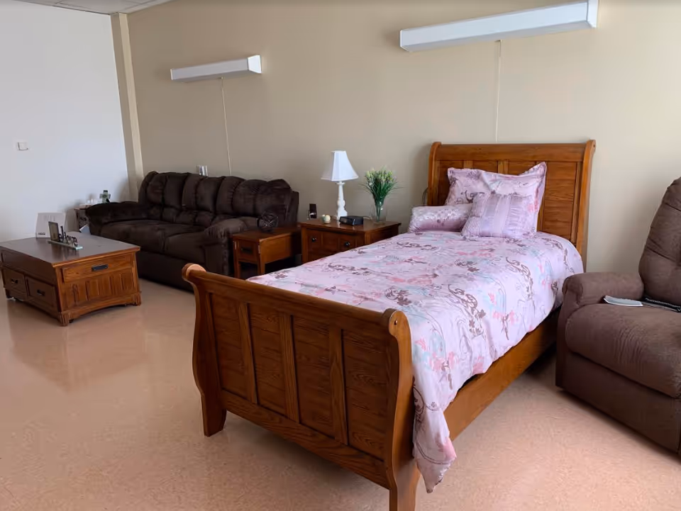 A cozy room featuring a wooden single bed with pink floral bedding and multiple pillows, a brown cushioned sofa, a brown recliner chair, a wooden coffee table, and a wooden nightstand with a white lamp and a small plant. The walls are beige, and the floor has a light brown finish.