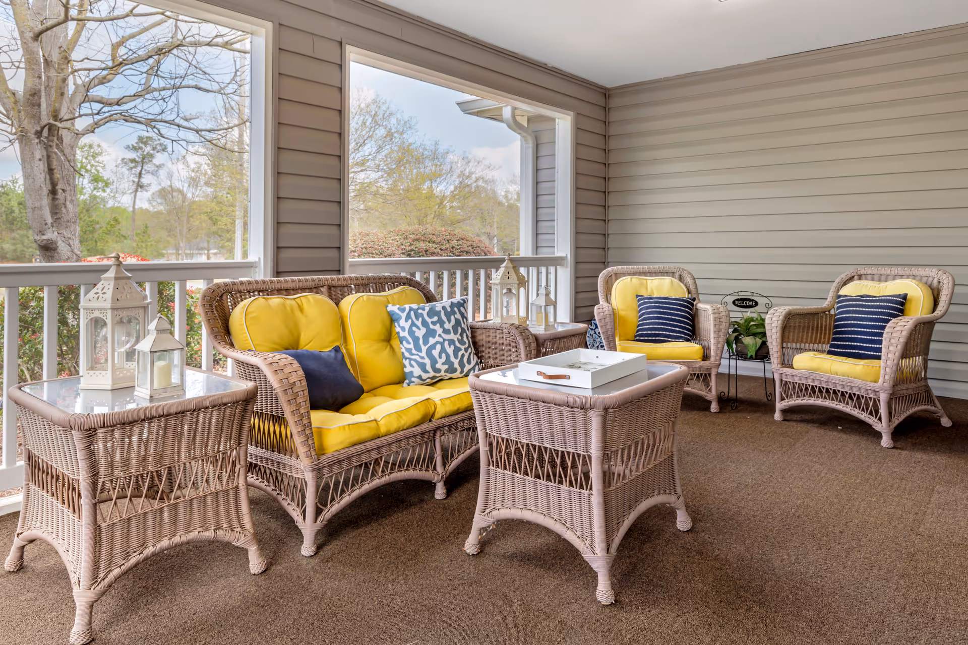 A cozy outdoor covered patio area with wicker furniture including a loveseat and three armchairs with yellow cushions and blue patterned pillows. There are two wicker side tables with decorative lanterns and a wicker coffee table with a white tray. The patio has beige siding walls and overlooks trees and greenery.