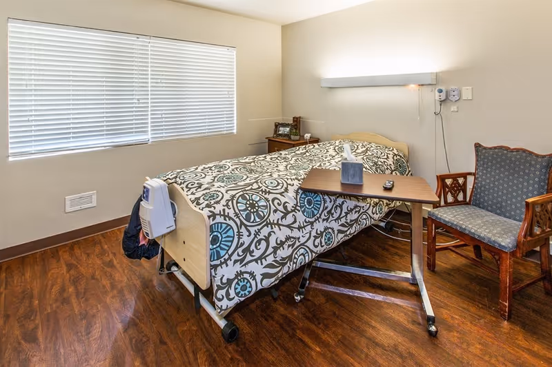 A small bedroom in a senior living facility with a single bed covered in a patterned bedspread. Next to the bed is a wooden bedside table with a photo frame and some items. A rolling overbed table with a tissue box and remote control is positioned over the bed. There is a cushioned wooden chair beside the bed. The room has wood flooring and a large window with closed blinds.