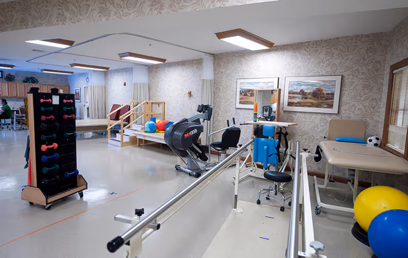 A physical therapy room in a senior living facility with exercise equipment including parallel bars, a stationary bike, therapy balls, dumbbells on a rack, and treatment tables. The room has floral wallpaper, framed landscape paintings on the wall, and bright overhead lighting.