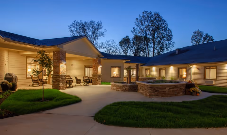 Outdoor courtyard area of a senior living facility at dusk with well-lit building exterior, patio seating, small trees, and landscaped grass areas.