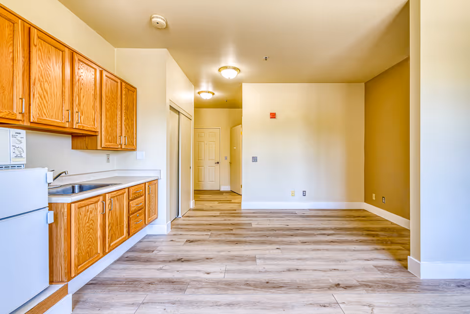 An empty interior space featuring a small kitchen area with wooden cabinets, a white refrigerator, and a microwave. The room has light-colored walls, wood-patterned flooring, and ceiling lights. There is a hallway leading to a closed door in the background.