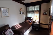 A small bedroom with a single bed against the wall, a wooden chair with a cushion, a nightstand with a lamp, and a window with sheer curtains letting in natural light. The walls have framed pictures and a small shelf with decorative items.