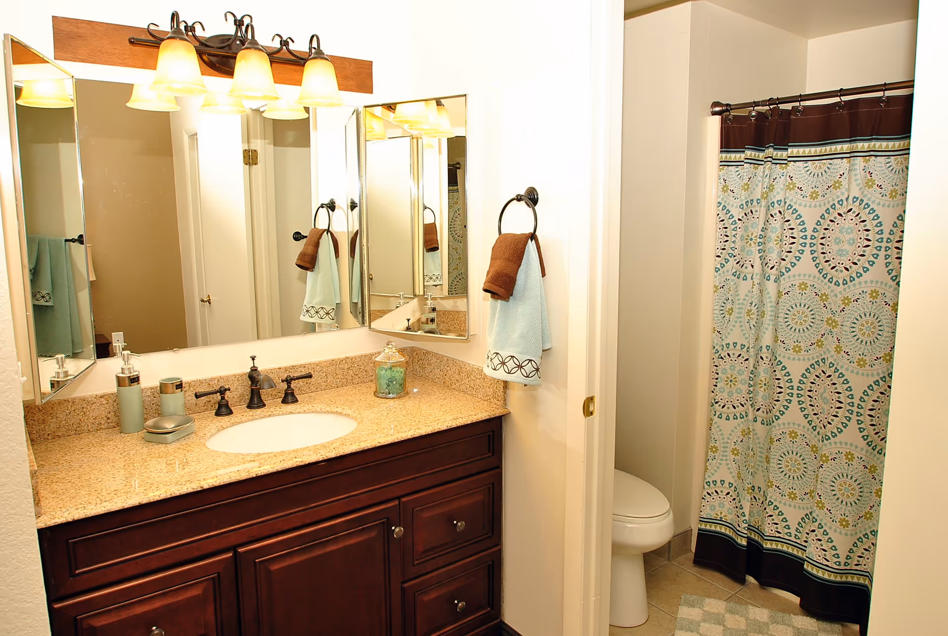 A bathroom with a granite countertop vanity featuring a sink, dark wood cabinets, and a large mirror with three light fixtures above. There are two towels hanging on a ring holder on the wall, one brown and one light blue. To the right, there is a toilet partially visible behind a door and a shower with a patterned curtain in shades of blue, green, and brown. The floor has beige tiles and a checkered bath mat.