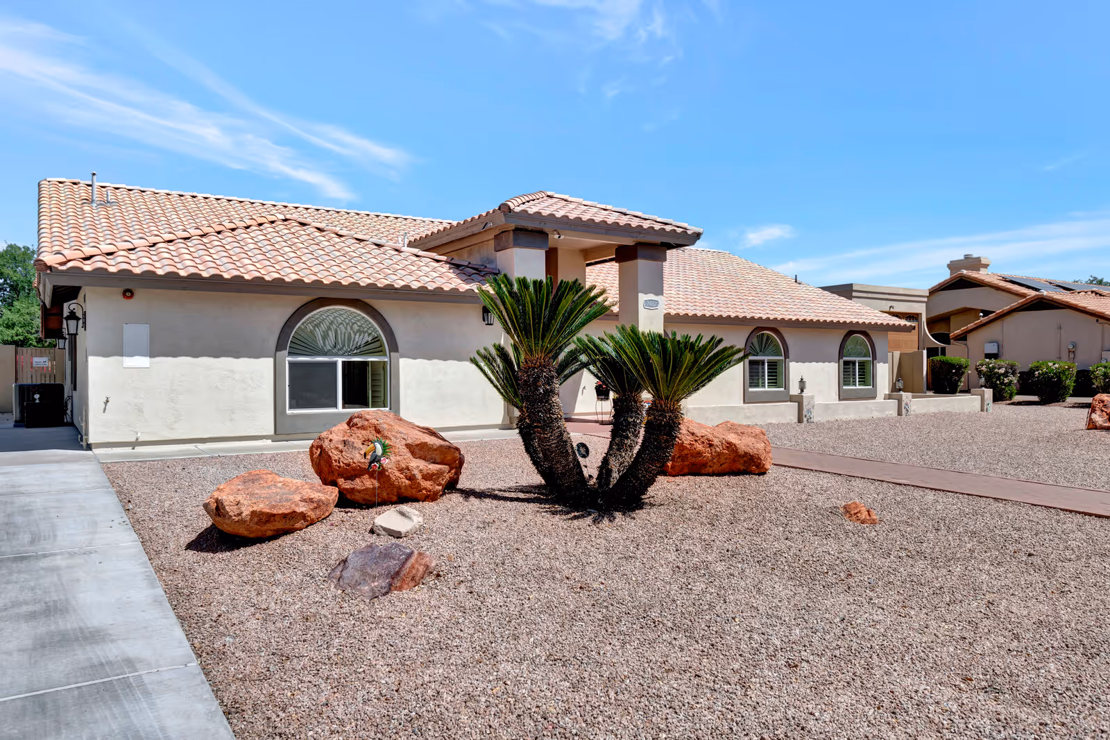 Exterior view of a single-story building with a tiled roof and arched windows, surrounded by a gravel yard with large rocks and a small palm plant under a clear blue sky.