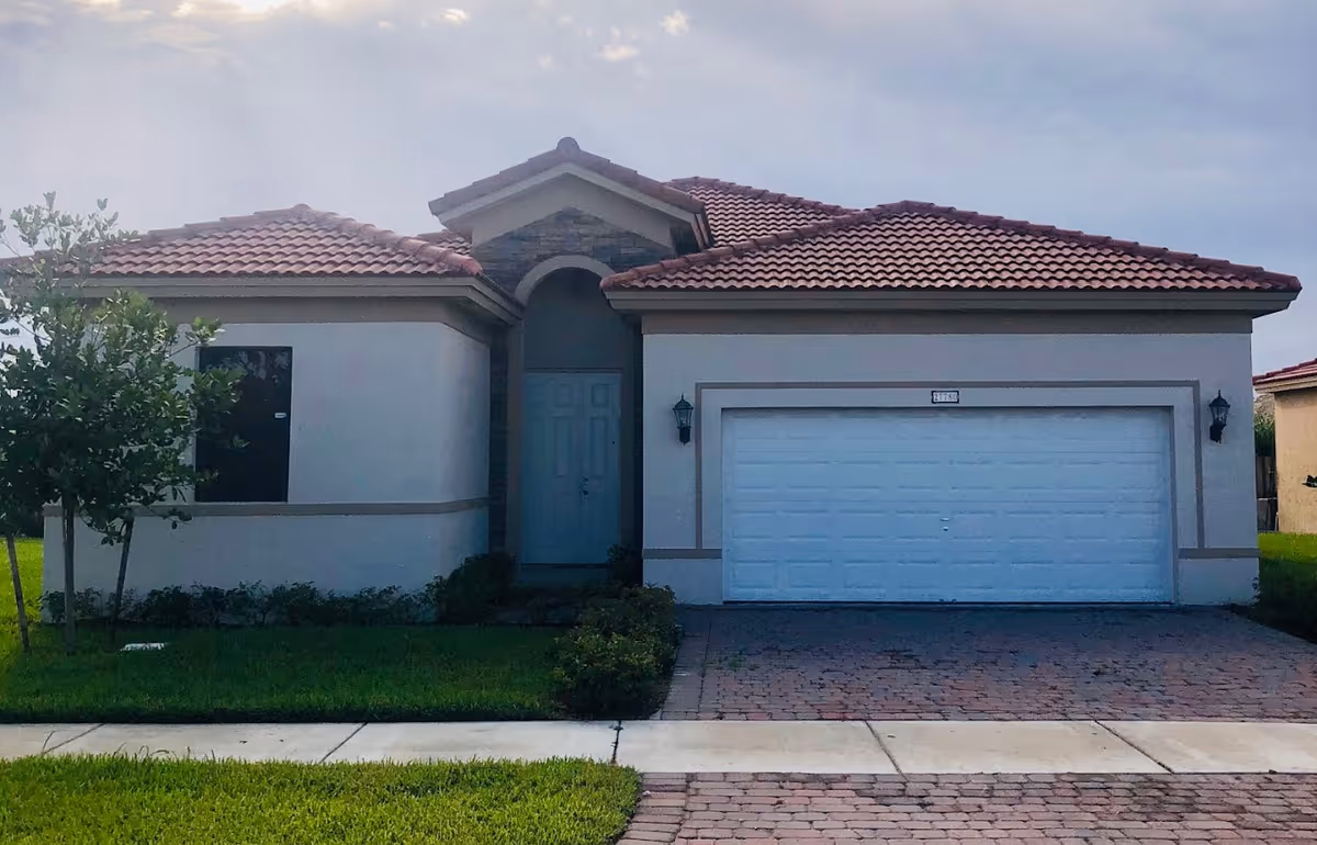 Front exterior view of a single-story house with a tiled roof, a double garage door, a central entrance door, and a small tree and shrubs in the front yard.