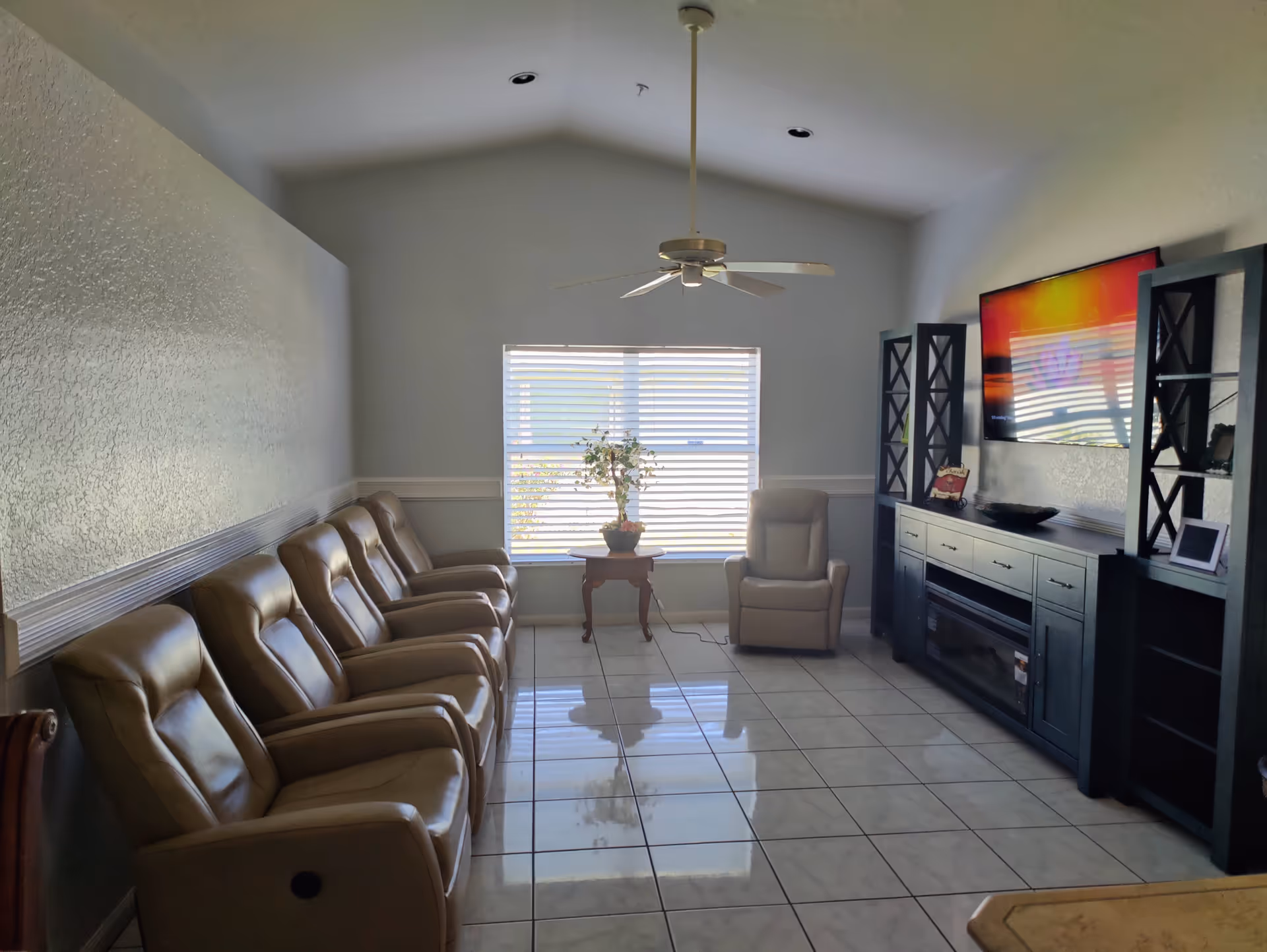 Sunlit common living room with a row of recliner chairs facing a TV and entertainment center, a window with blinds and a small table with a plant.