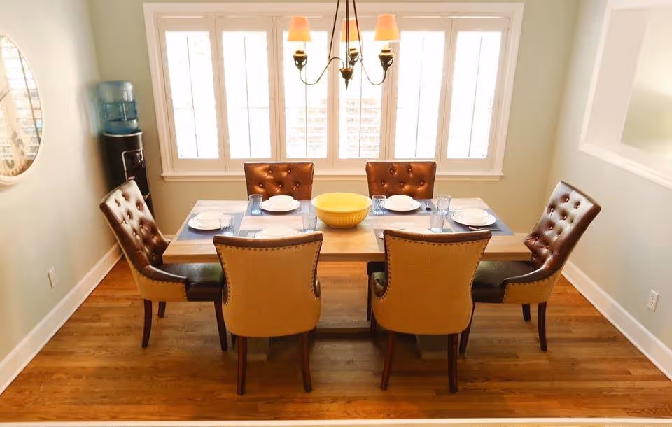 A dining room with a wooden table set for six people. The table has six chairs with brown leather upholstery and beige backs. There are plates, glasses, and a yellow bowl on the table. A chandelier with five lamps hangs above the table. Large windows with white shutters are in the background, and a water cooler is visible in the corner.