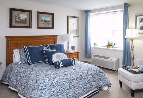 A neatly made bed with a blue and white patterned comforter and multiple blue pillows in a senior living bedroom. The room has a wooden headboard, a nightstand with a lamp and a framed photo, a window with blue curtains, a heating/cooling unit below the window, and a comfortable armchair with a small table holding books and a cup.