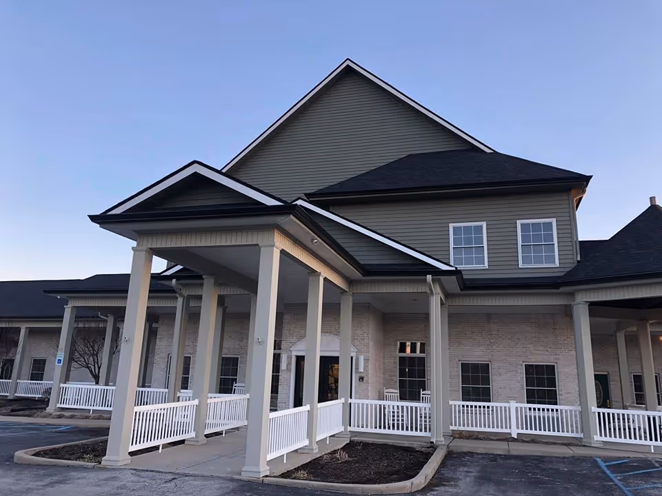 Front entrance of a multi-story senior living building with a covered portico supported by columns, white railings, and adjacent parking.
