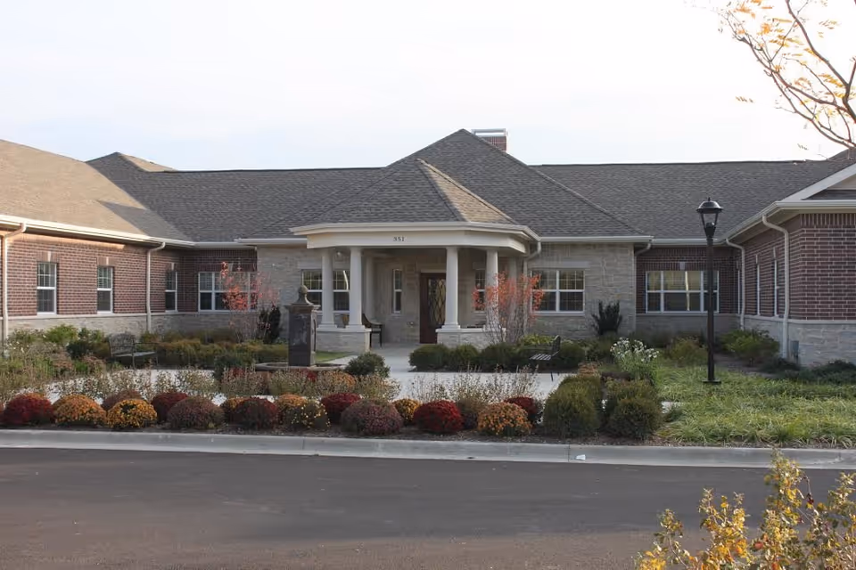 Front entrance of a single-story brick and stone senior living building with columns, landscaping, benches, and a driveway.